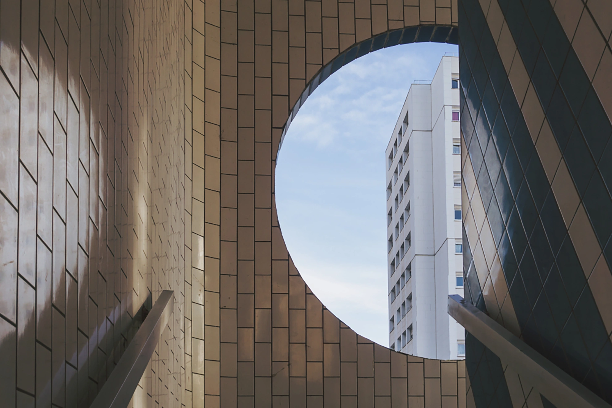 A circular opening in a tiled structure framing a tall white building against a partly cloudy sky