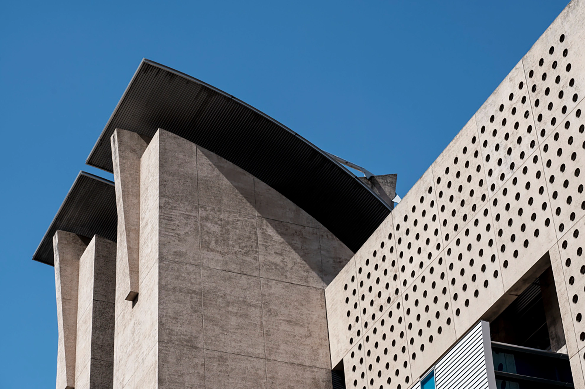 Modern concrete building with perforated facade and angular roof against a clear blue sky