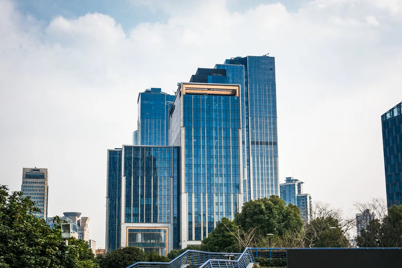 Tall glass skyscrapers with blue-tinted windows, surrounded by trees and a pedestrian bridge, under a partly cloudy sky