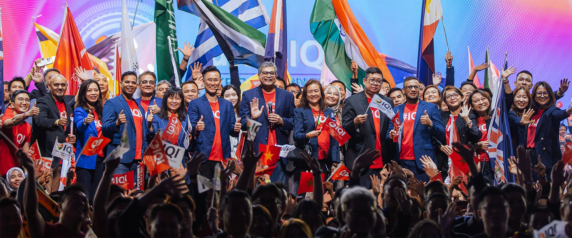 A large group of people on stage at an international event, smiling and waving while holding various national flags and IQI banners.